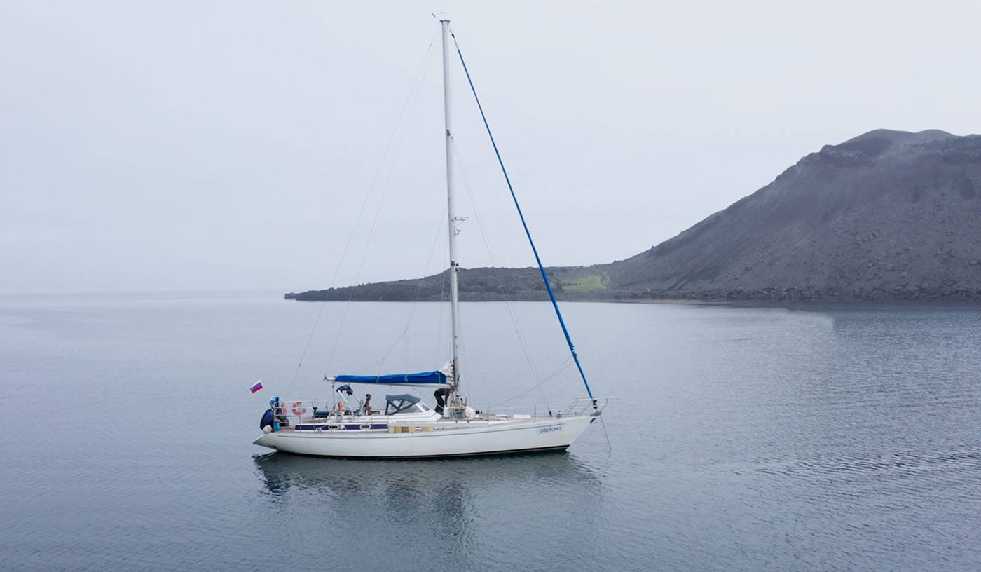 Парусная яхта Stella Maris на якоре в тихой бухте у тёмного склона, пасмурно