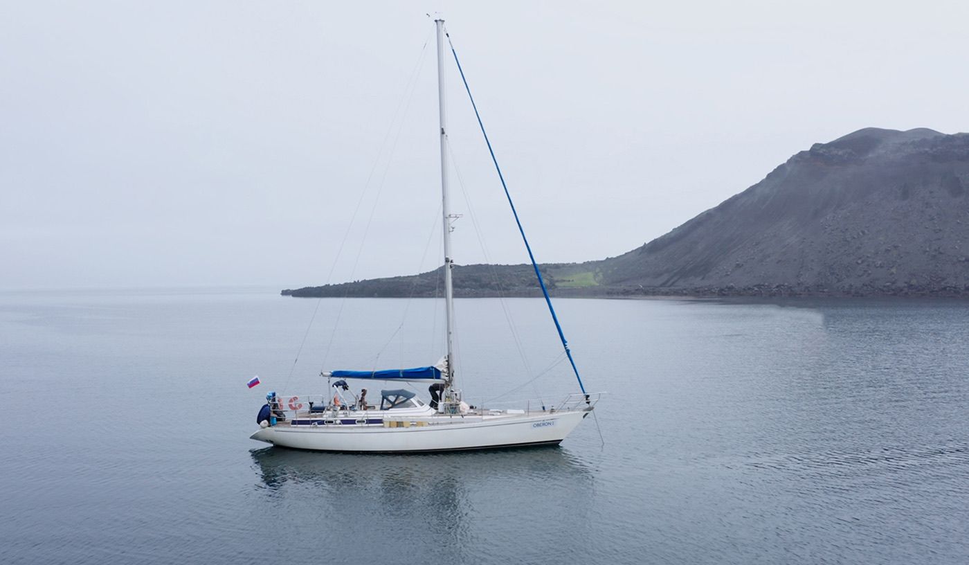 Парусная яхта Stella Maris на якоре в тихой бухте у тёмного склона, пасмурно