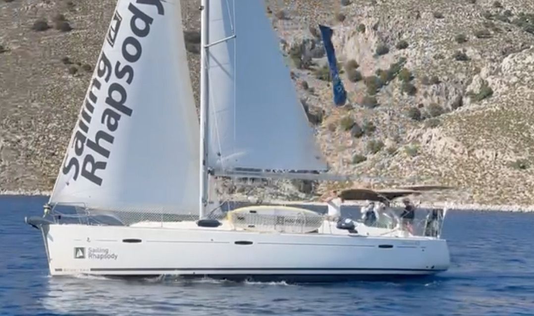 Rhapsody идёт под гротом и генуей вдоль гористого берега, на гроте надпись Sailing Rhapsody